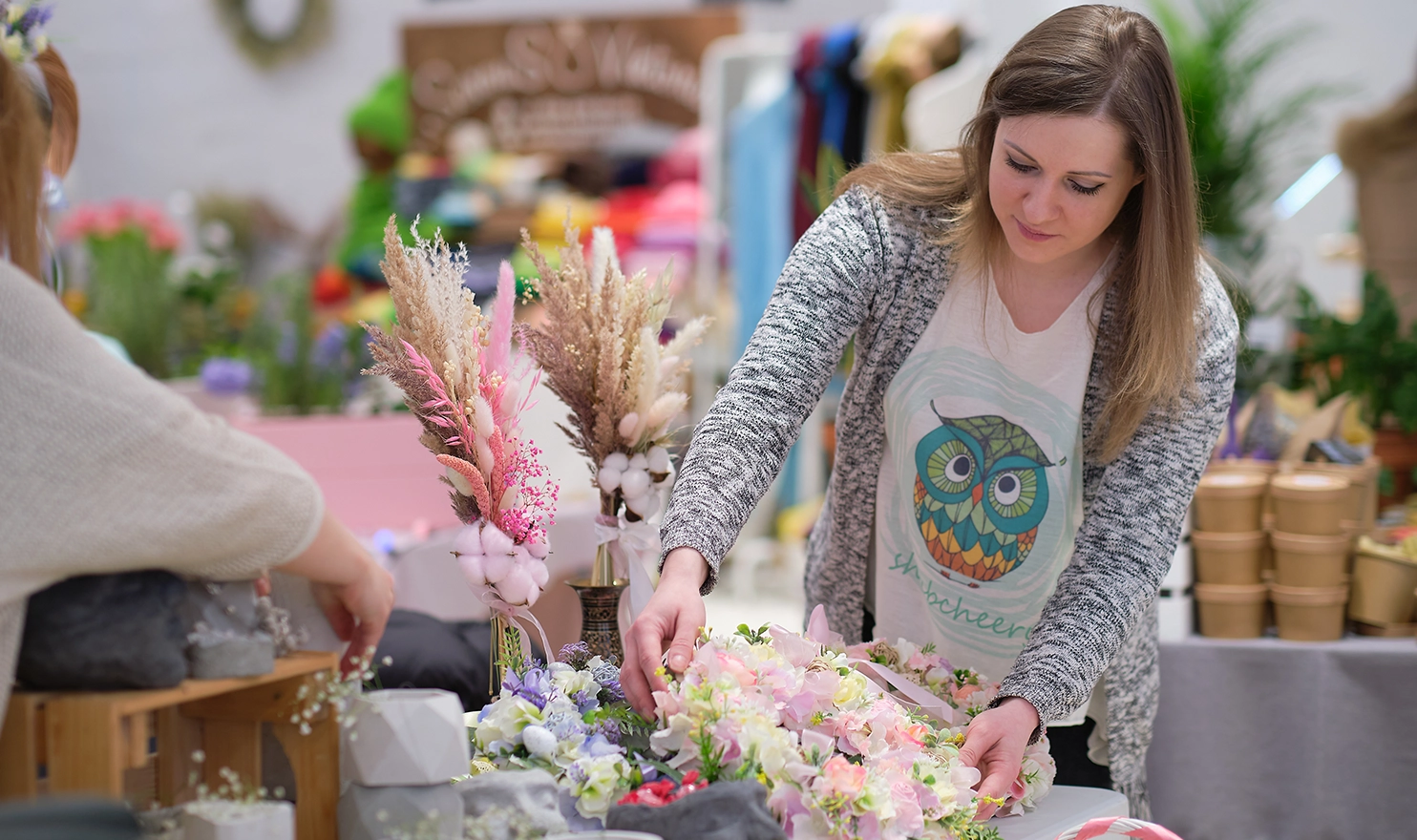 A crafter sets up her handmade floral wreaths in her booth at an indoor craft fair.