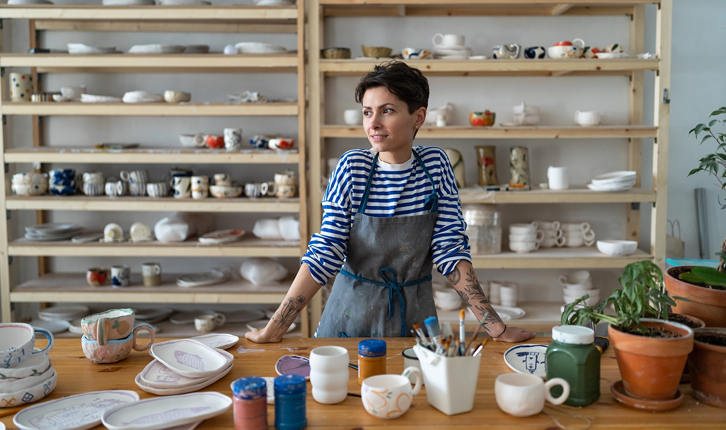 A female potter is standing in her studio with pieces in front of her and behind her on shelves.