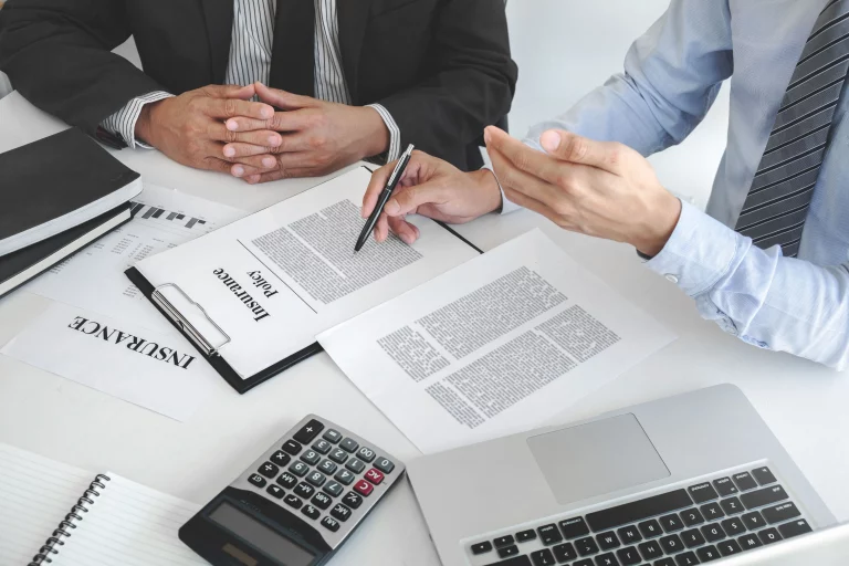 Two business professionals in formal attire are seated at a table, discussing an insurance policy document. One person is holding a pen, pointing at the policy, while the other listens attentively with folded hands.