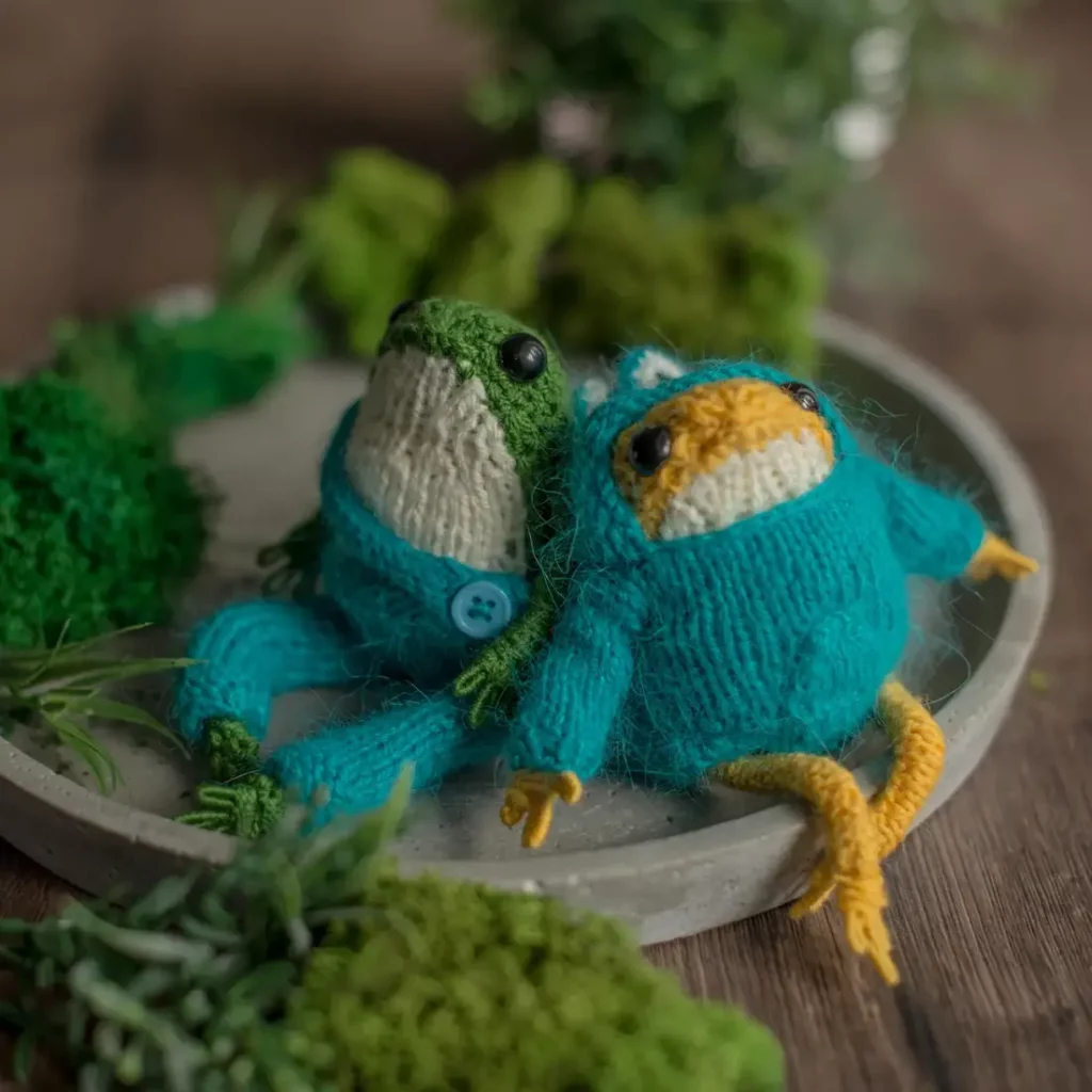 A pair of crochet frogs wearing blue outfits pose on a gray dish with green moss surrounding them.