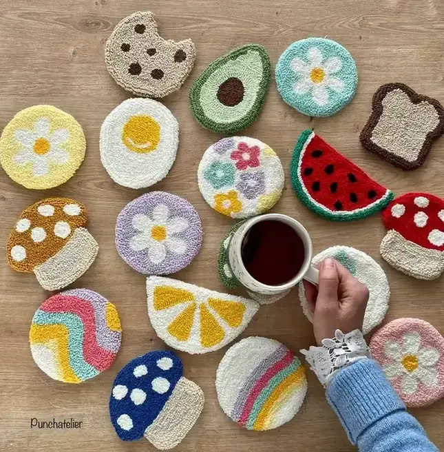 A flat lay of colorful punch needle coasters, with designs including an egg, watermelon, mushrooms, and flowers, on a wooden table with a hand holding a mug of tea above one of the coasters.