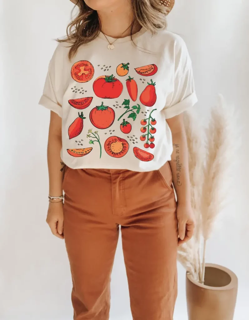 A female model wears a cream-and-red tomato-print crewneck T-shirt and brown pants in front of a brown vase with pampas grass.