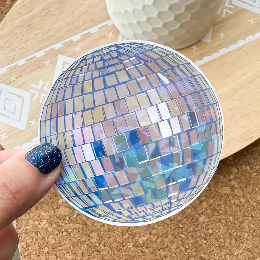 A blue and silvertone disco ball sticker being held by a hand above a wooden table.