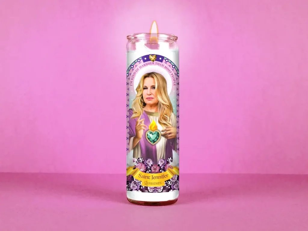 A white candle with a label that depicts Jennifer Coolidge illustrated as a saint with text that reads, "I'm kind of harsher than most people" and "Saint Jennifer, Lit Friends."