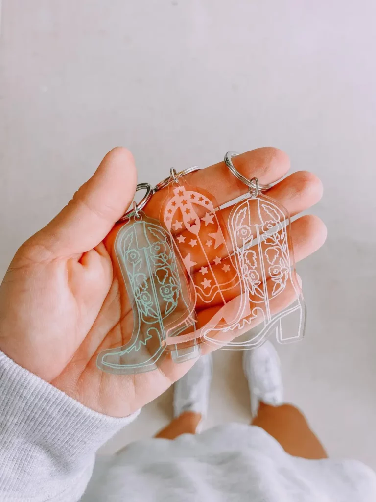 A hand holds three clear cowboy boot key chains with star and floral prints.