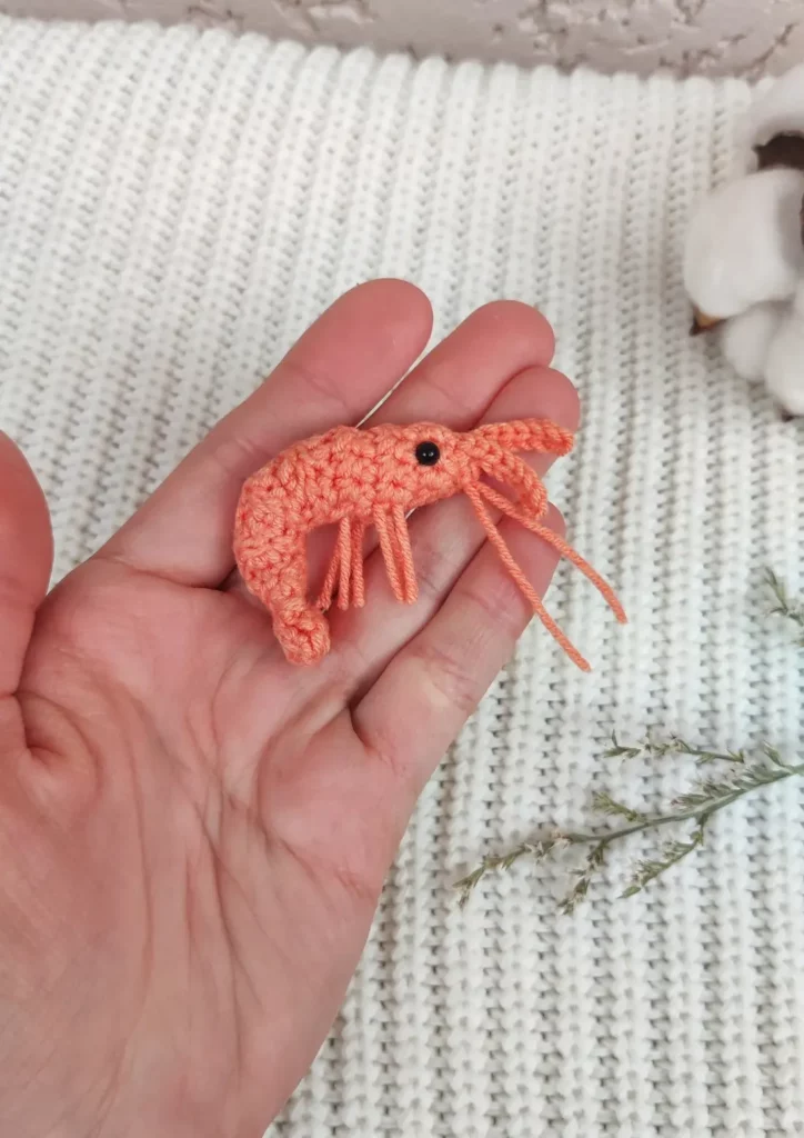 A hand holding a miniature pink crochet shrimp toy.