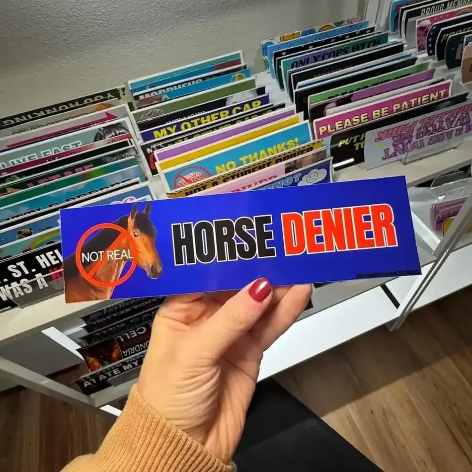 A blue and red bumper sticker that displays an image of a horse with the text, "Horse denier" and "Not real" being held in front of a collection of bumper stickers.