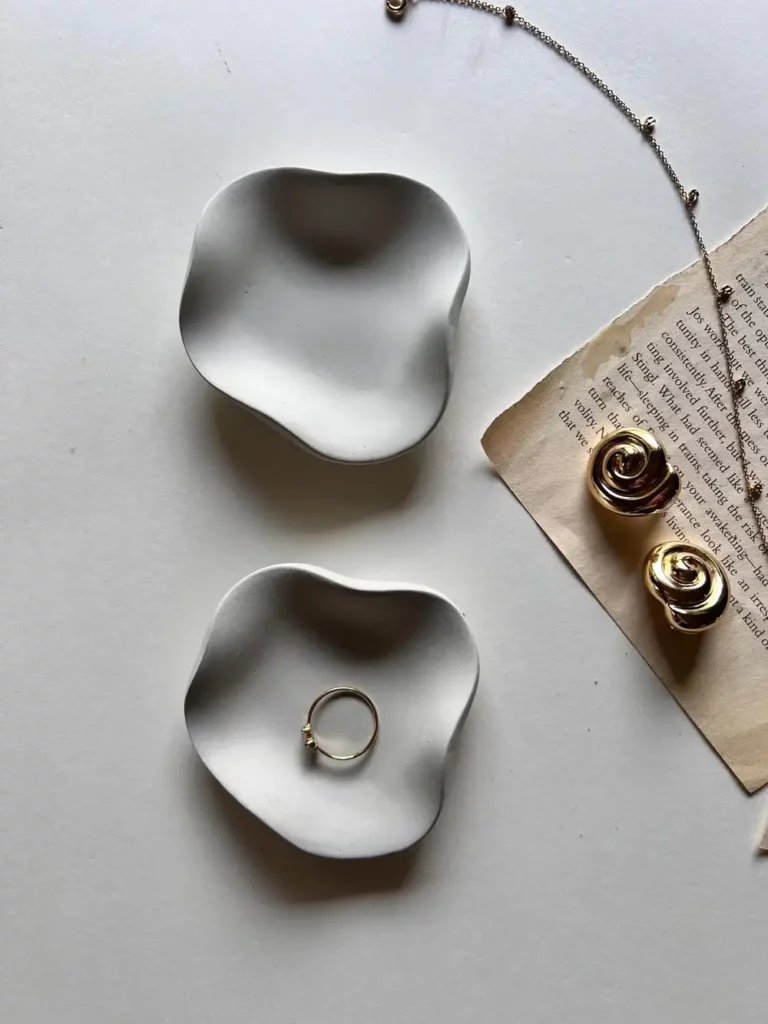 A set of white minimalist, organic-shaped jewelry trays, a worn book page, and goldtone jewelry laid out on a white table.