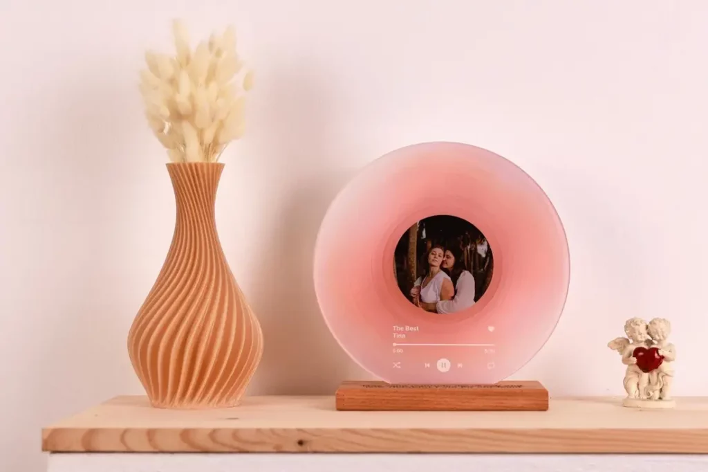 A pink personalized record decor with a couple's photo at its center is displayed on a wooden stand along with a curved vase with pampas grass and a cherub figurine.