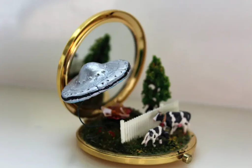 A goldtone compact mirror diorama depicting a farm scene with cows, a tree, a white fence, and a silver UFO hovering above.